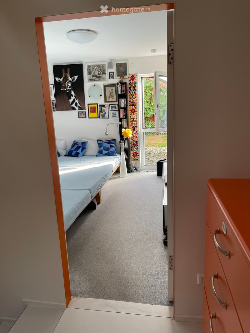 Modern bedroom with a bed, pillows, bookshelf, vase, lamp, and picture frames on the wall. The room has a carpeted floor and a large window with a view of greenery.
