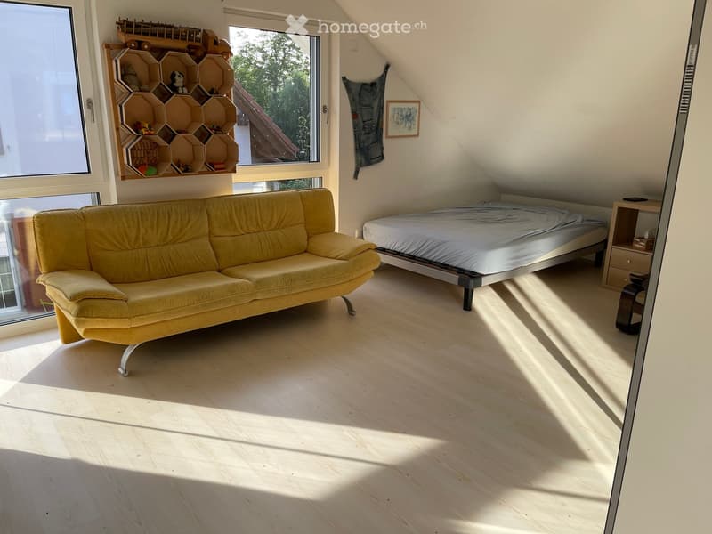 Bright, multi-functional room with a sofa, bed, nightstand, and a wooden shelf with toys and decor. Large windows allow natural light to fill the space.