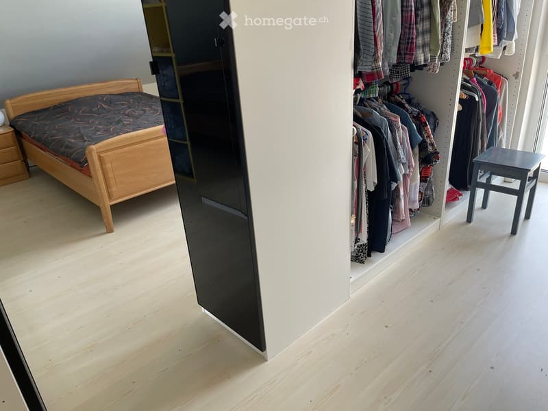 A modern bedroom with a wooden bed, nightstand, and an open wardrobe filled with clothes. The floor is light wooden, and a small black stool is placed near the wardrobe.