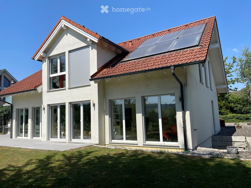 Two-story house with red-tiled roof, large windows, solar panels, and a well-maintained lawn.