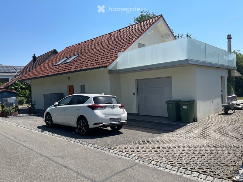 Two-story house with red-tiled roof, white walls, garage, and a modern glass balcony on the upper level. A white car is parked in front of the house.