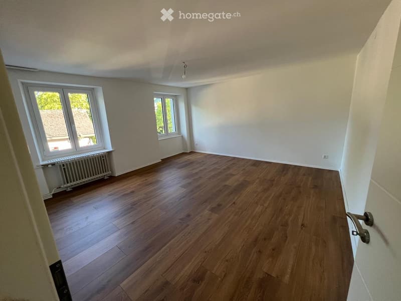 Empty room with wooden flooring, white walls, and large windows offering a view of a roof and greenery outside.
