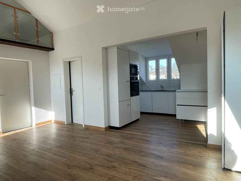 Bright, unfurnished room with wooden flooring, white walls, and large windows. Features a modern kitchen area with white cabinets and stainless steel appliances visible in the background.