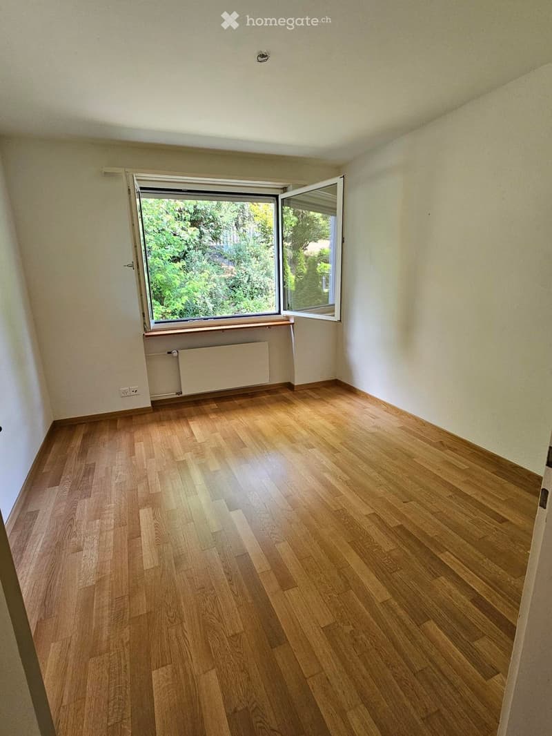 Empty room with light wooden flooring, white walls, and a large window with open shutters showing greenery outside.