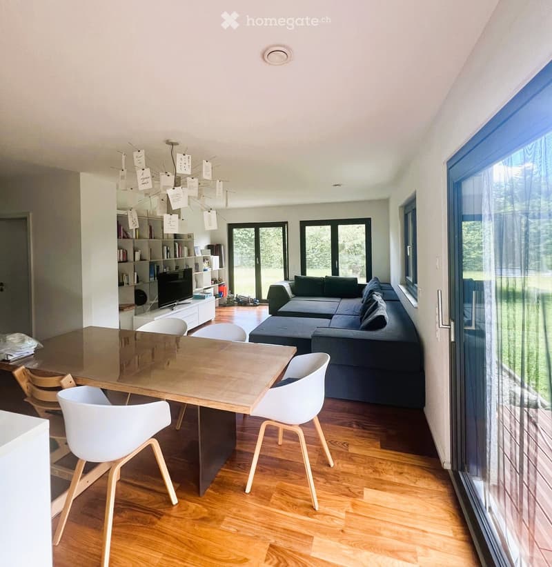 Modern living and dining area with a dark grey sectional sofa, a wooden dining table with four white chairs, a television on a white stand, and a bookshelf with books and decorative items.