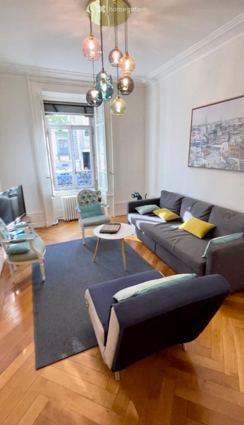 Modern living room with wooden flooring, large window, grey sofa, coffee table, armchairs, ottoman, floor lamp, and a television.