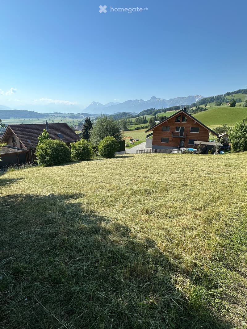 Two-story wooden house with a red-tiled roof, surrounded by green fields and mountains in the background.