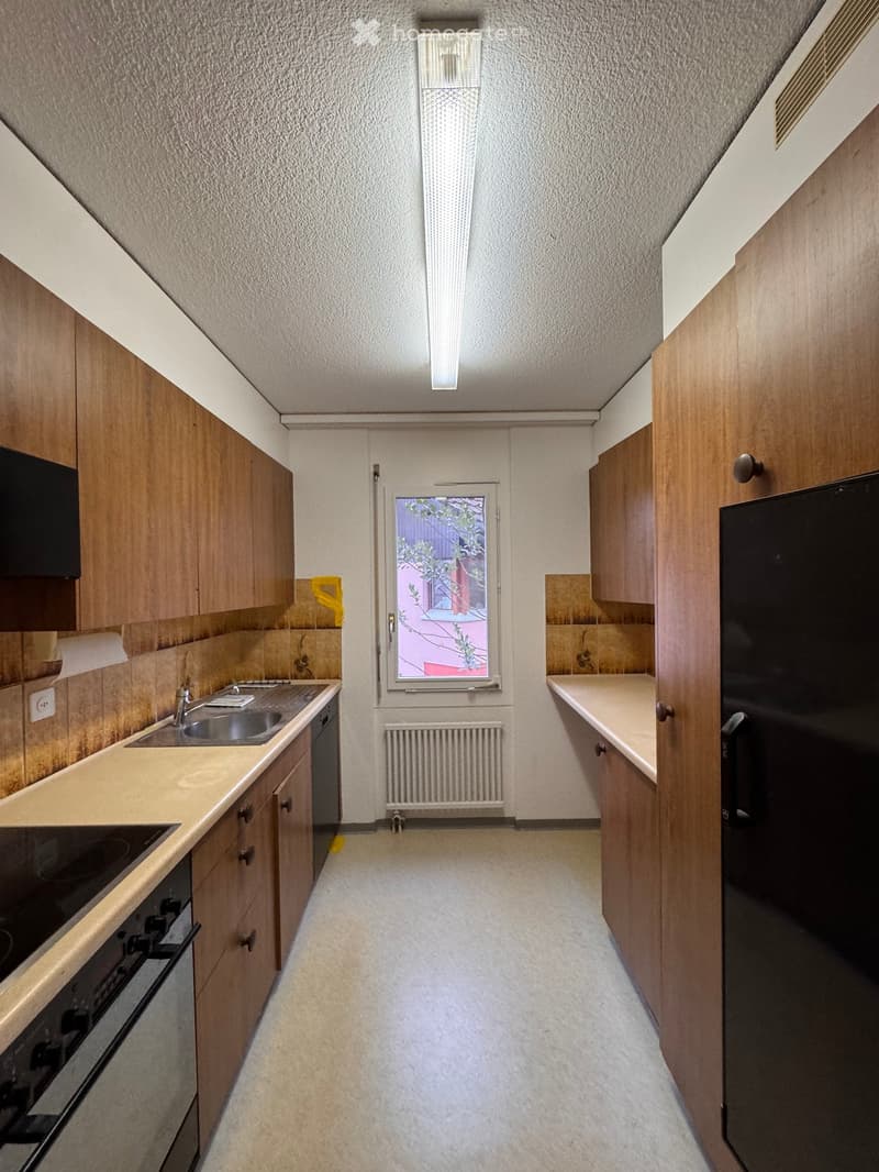 Compact kitchen with wooden cabinets, a sink, oven, and refrigerator. Features a window with a view of a neighboring building.