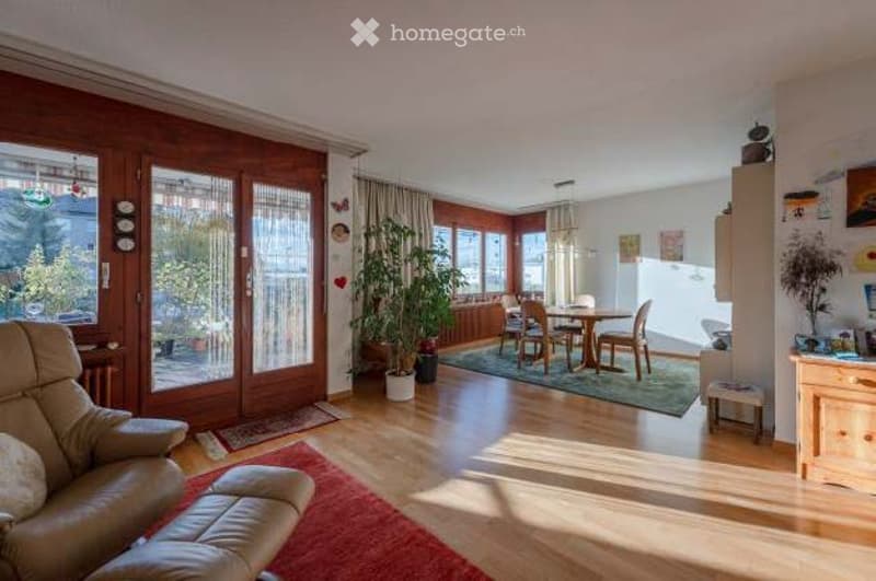 Bright living and dining area with large windows, wooden flooring, a leather sofa, ottoman, dining table with chairs, potted plants, and a cabinet.