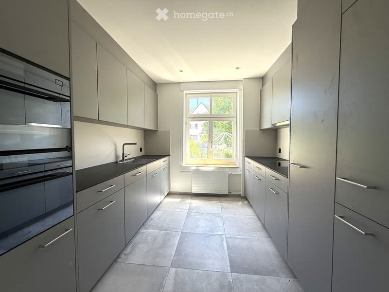 Modern kitchen with grey cabinets, a central island, and a large window overlooking a garden.
