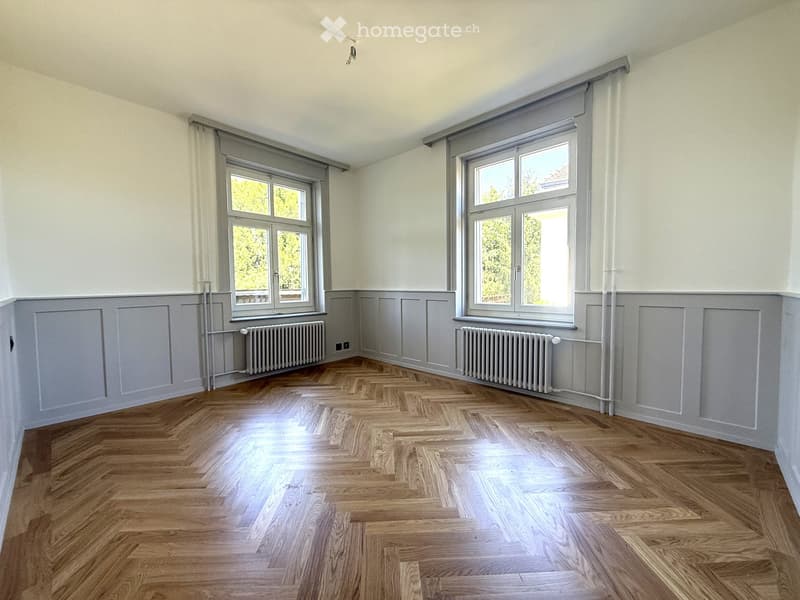 Empty room with wooden herringbone flooring, white walls, and two windows with grey frames. Radiators are present under each window.