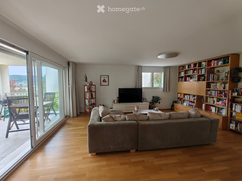 Modern living room with wooden flooring, large sliding glass doors leading to a balcony, a grey sectional sofa, coffee table, bookshelf, and television.