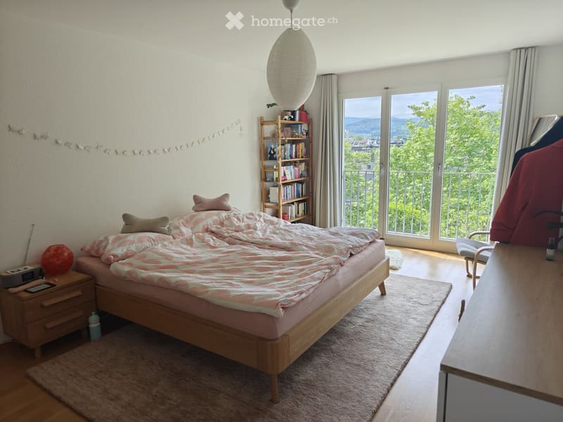 Cozy bedroom with a wooden bed, nightstand, bookshelf, and large windows offering a scenic view. Includes a chair, table, and decorative lamp.