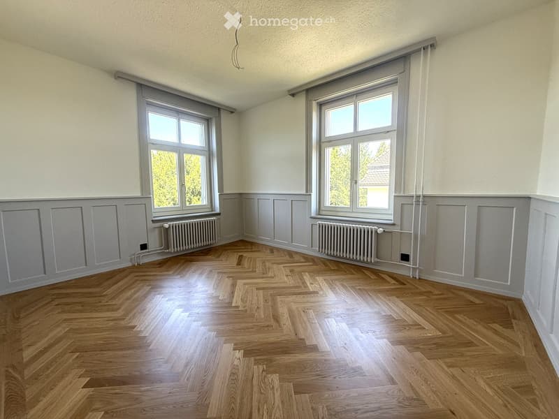 Empty room with wooden herringbone flooring, white walls, and two large windows with grey frames. Radiators are present under each window.