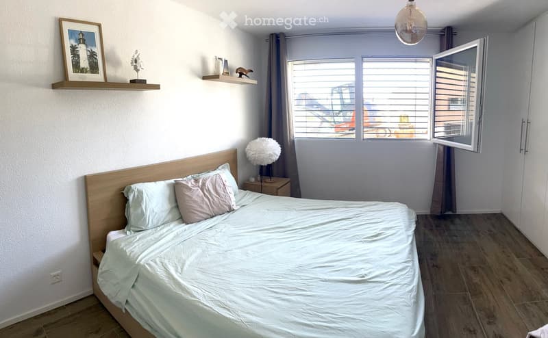 Modern bedroom with a wooden bed, light-colored bedding, a nightstand with a lamp, and a shelf with decorative items and a picture frame. The room features large windows with blinds and a view of the outside.