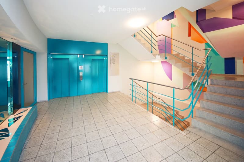Modern hallway with a staircase featuring colorful panels and a teal railing, an elevator on the left, and a tiled floor.