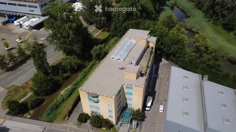 Aerial view of a modern multi-story building with a flat roof, surrounded by parking areas, greenery, and nearby industrial structures.