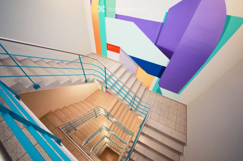 Staircase with colorful, abstract wall art and modern railings, leading to an open area with wooden flooring and tiled steps.