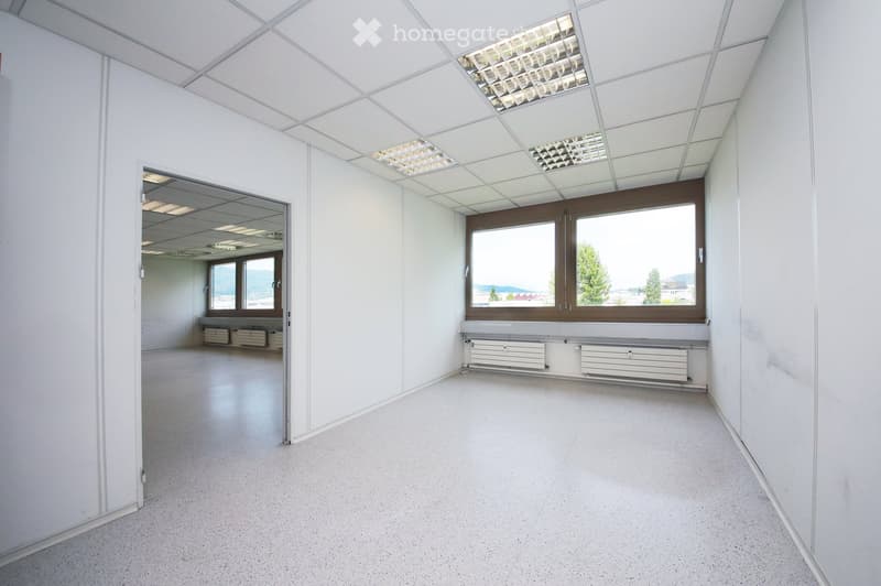 Empty room with white walls, a large window with a view, and a radiator below it. The room has a smooth, light-colored floor and a ceiling with recessed lighting.