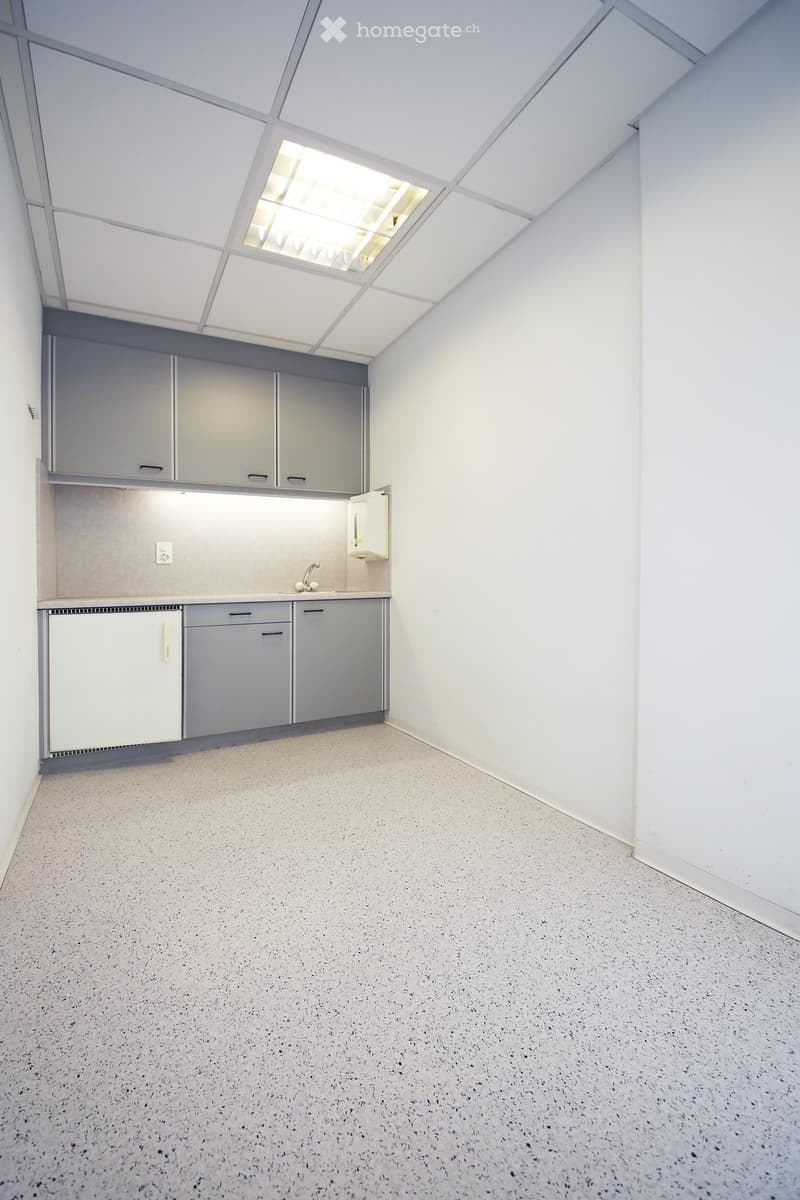 Empty room with grey cabinets, sink, and a small refrigerator. The ceiling has a skylight, and the floor is a speckled concrete surface.