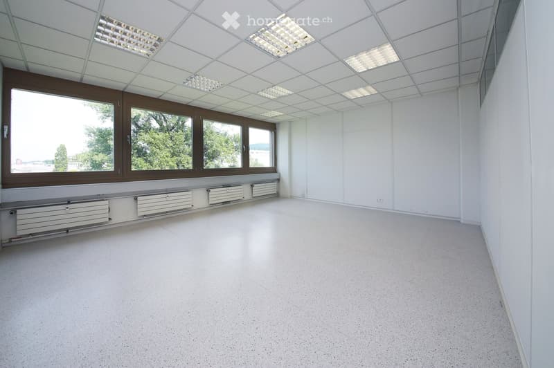 Empty room with white walls, a white ceiling with recessed lighting, large windows with brown frames, and white radiators along the wall.