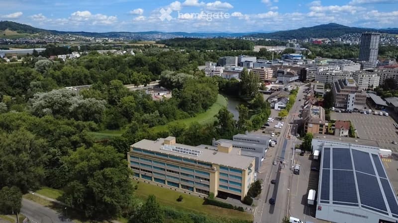 Aerial view of a cityscape with a mix of commercial buildings, parking lots, and green spaces, surrounded by trees and a river.