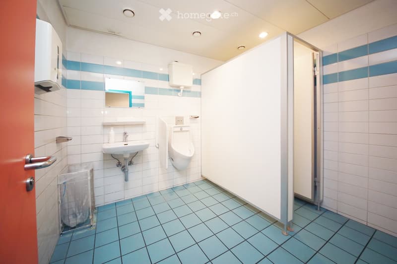 Clean public restroom with white and blue tiled walls, a sink, urinal, mirror, toilet paper dispenser, and a trash bin. The floor is covered with light blue tiles.
