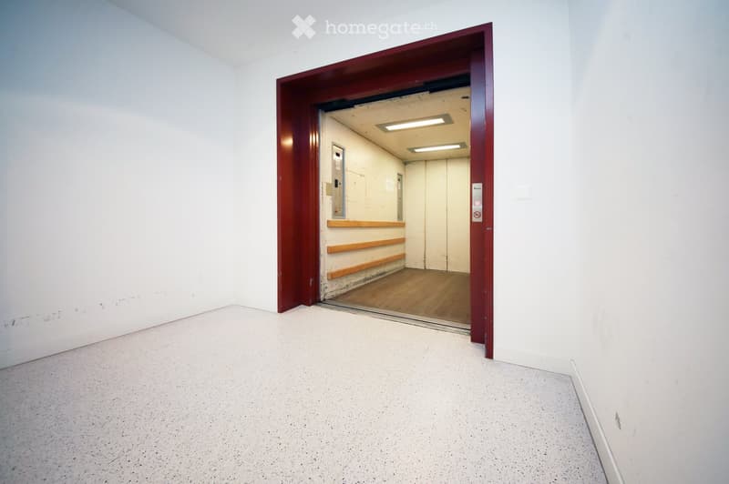 Empty room with white walls and a concrete floor, featuring an open doorway leading to another room with wooden flooring and a visible elevator shaft.