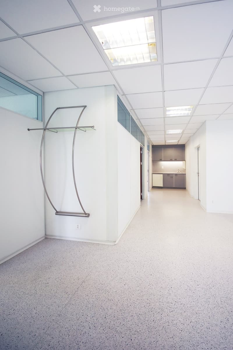 Open, modern corridor with white walls, a sleek floor lamp, and a small kitchenette area visible at the end.