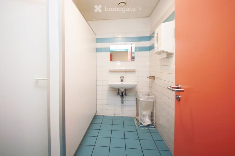 Clean bathroom with white tiled walls, blue tiled floor, a white sink, a mirror, a paper towel dispenser, and a small trash bin.