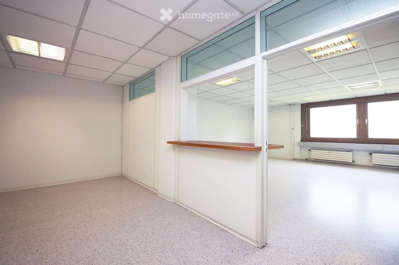 Empty room with white walls, carpeted floor, and large windows. Features include a wooden counter and radiators along the wall.