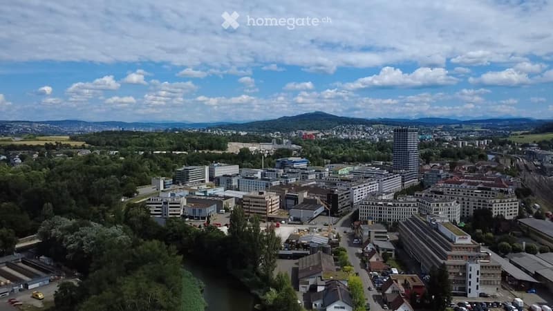 Aerial view of a cityscape with a mix of residential and commercial buildings, surrounded by greenery and a river running through the area.