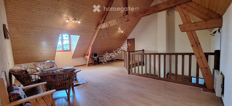 Spacious living area with wooden beams, exposed wooden ceiling, large window, and a balcony overlooking the area. Features include a sofa, chairs, a small table, a computer on a desk, and warm lighting.
