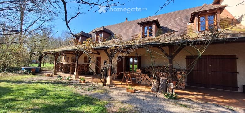 A two-story house with a tiled roof, wooden beams, and a covered porch area. The house features a mix of stone and wood exterior, with a well-maintained garden and stacks of firewood nearby.