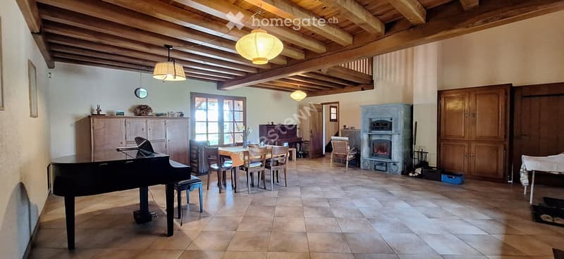 A spacious room with exposed wooden beams, a grand piano, a dining table with chairs, a fireplace, and a cabinet.