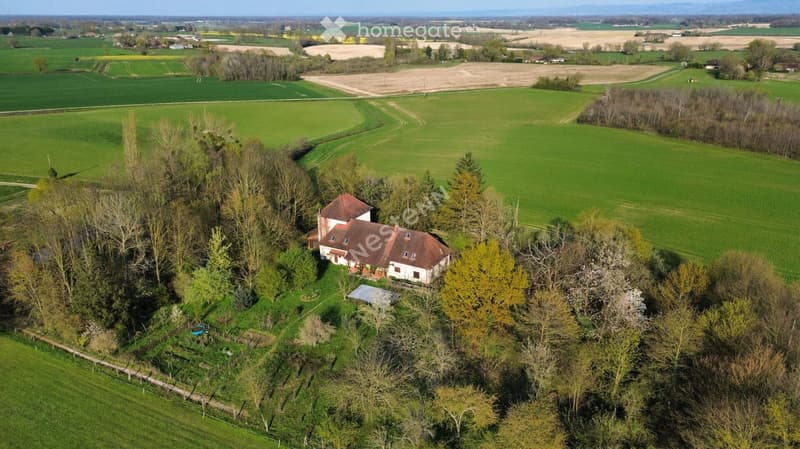 Aerial view of a single-story house with a red-tiled roof, surrounded by trees and greenery, located in a rural area with open fields in the background.