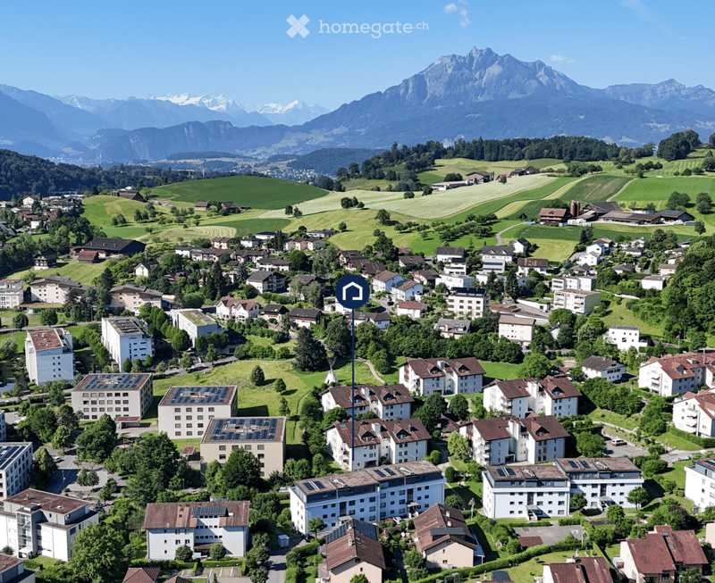 Aerial view of a town nestled in a valley with surrounding mountains, featuring numerous residential buildings, green fields, and scattered trees.