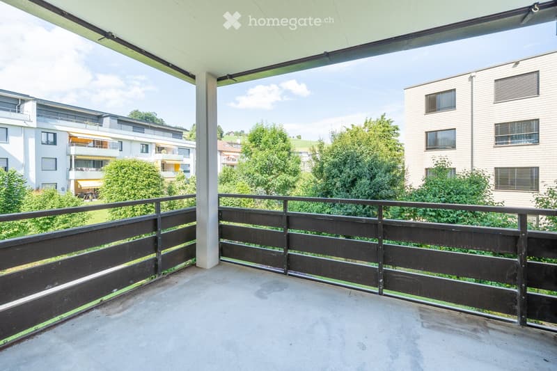 Balcony with wooden railing, concrete floor, and view of surrounding greenery and buildings.