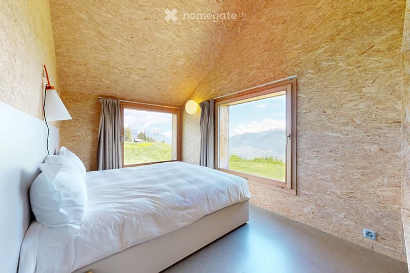 Modern bedroom with a large bed, white bedding, and large windows offering a scenic mountain view. The room features wooden panel walls and a minimalist design.