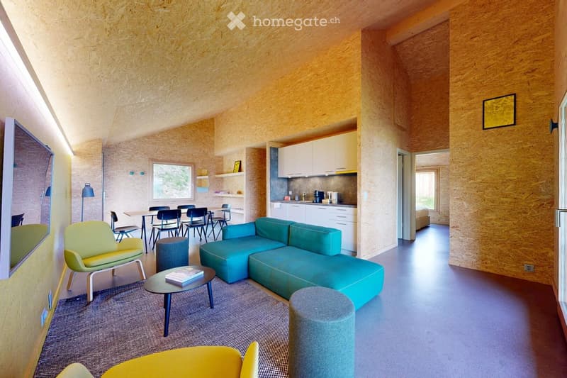 Modern living and dining area with a teal sectional sofa, coffee table, yellow armchair, dining table with chairs, and a small kitchen area with white cabinets and appliances.
