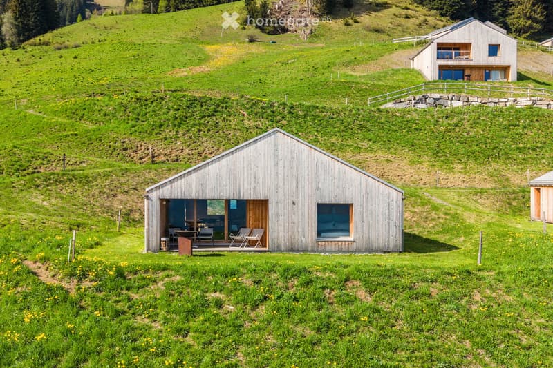 A modern wooden house situated on a grassy hillside, featuring large windows and an open porch area with outdoor furniture.