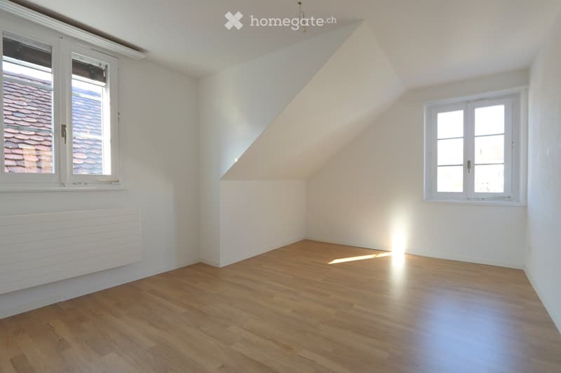 Empty attic room with wooden flooring, white walls, and two windows offering views of the roof.