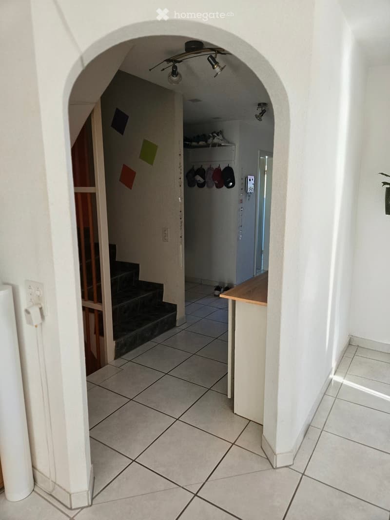 A clean, tiled hallway with an arched entryway leading to a staircase and a small storage area with shelves and hanging hooks.