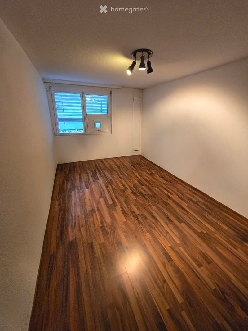 Empty room with wooden flooring, white walls, and a small window with blinds. A ceiling light fixture is visible.