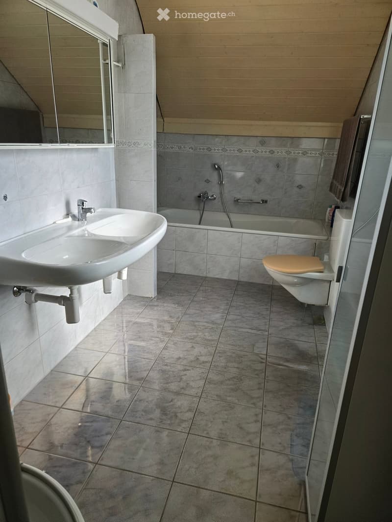Modern bathroom with a white sink, toilet, bathtub, and mirror. The walls are tiled with grey tiles and the floor is covered with large, light grey tiles.