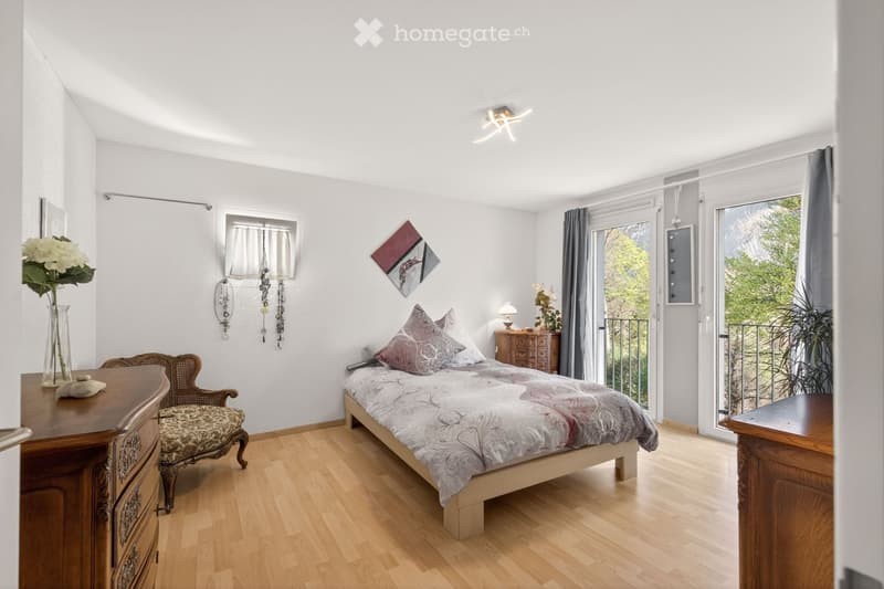 Cozy bedroom with wooden flooring, a large bed with patterned bedding, a nightstand with a lamp, a wooden dresser, a decorative chair, and a vase with flowers on a side table. The room features large windows with curtains and a view of greenery outside.