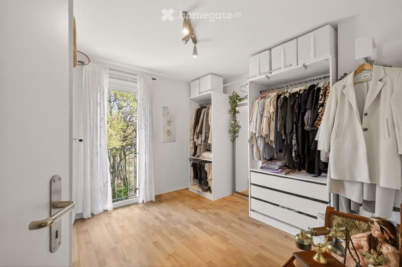 A well-organized walk-in closet with built-in wardrobes, drawers, and shelves. The space features wooden flooring, white walls, and a large window with white curtains. A small table with a vintage telephone and a doll is placed near the door.