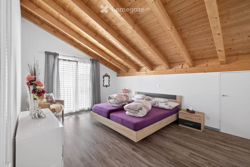 Cozy bedroom with exposed wooden beams, two single beds with purple bedding, a nightstand, a mirror, a small table with a vase of flowers, and a chair with a fur throw.
