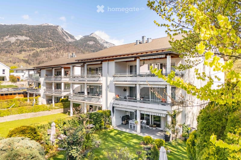 A large, modern multi-story house with white exterior and multiple balconies, surrounded by a well-maintained garden and lush greenery, with a scenic mountain view in the background.