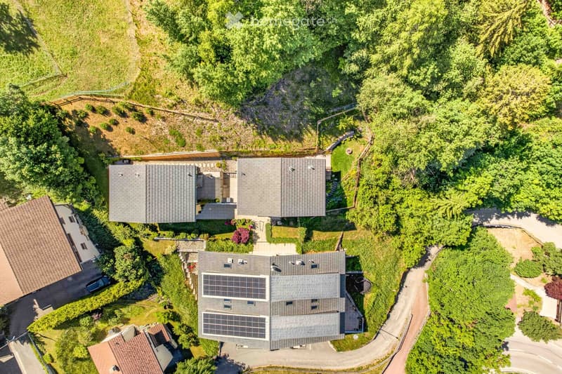 Aerial view of a modern house with solar panels on the roof, surrounded by lush greenery and trees. The house features multiple sections and is situated on a raised ground floor with a driveway leading to it.